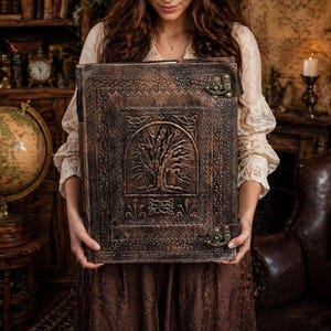 May include: A large, brown leather-bound journal with an embossed tree design on the cover. The journal has decorative metal clasps and intricate border details. The book is held in front of a person, suggesting a focus on craftsmanship and a vintage aesthetic.