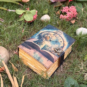 May include: A rectangular journal with a printed cover featuring a clock face and an open book illustration. The journal has thick, deckled-edge pages with a yellowish hue and is secured by a dark metal latch attached to a black strap on the side.