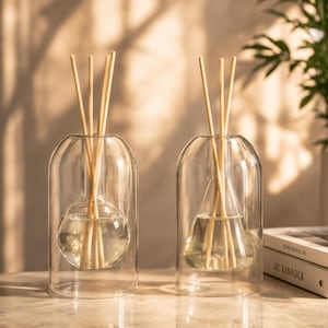 Peut inclure: Deux diffuseurs de parfum à bâtonnets en verre transparent avec une conception à double paroi. L'un possède un récipient intérieur sphérique, l'autre un récipient conique. Ils contiennent un liquide clair et des bâtonnets en bois clair.