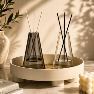 Peut inclure: Deux diffuseurs de parfum coniques en verre reposent sur un plateau rond de couleur crème à pieds courts. L'un est en verre gris foncé avec trois bâtonnets blancs, l'autre en verre transparent avec trois bâtonnets noirs, contenant de l'huile.