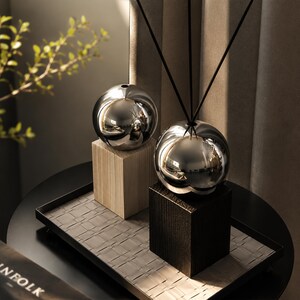 May include: Two spherical silver-toned diffuser vessels with reflective surfaces, each mounted on a square wooden base. One base is light-colored wood, and the other is dark-colored wood. Thin black reeds extend upward from the top of each sphere.