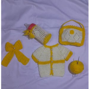 May include: A collection of handmade crochet baby items in cream and yellow. The set includes a bottle cover, a bow, a purse, a sweater, and a ball of yarn. The items are arranged on a white surface.