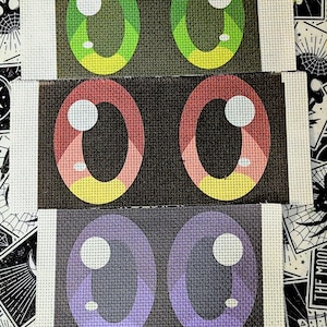 May include: Three sets of cartoon eye designs on canvas. The top set features green and yellow eyes, the middle set has pink and yellow eyes, and the bottom set has purple eyes. Each eye has a white highlight.