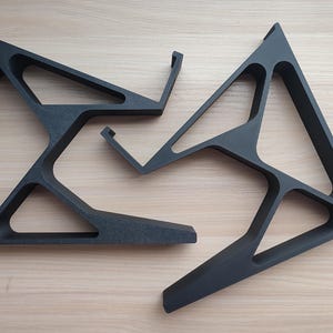Könnte beinhalten: Zwei schwarze Laptop-Ständer mit matter Oberfläche und offenem, geometrischem, dreieckigem Rahmendesign. Jeder Ständer verfügt über eine breite Basis, einen geneigten Stützarm und eine kleine Hakenlippe oben, um ein Gerät sicher zu halten.