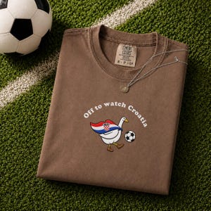K&ouml;nnte beinhalten: Ein braunes T-Shirt mit kurzen &Auml;rmeln und einer Grafik einer wei&szlig;en Gans, die ein Cape in den Farben der kroatischen Flagge tr&auml;gt und einen Fu&szlig;ball kickt. Der Text &uuml;ber der Gans lautet "Off to watch Croatia".