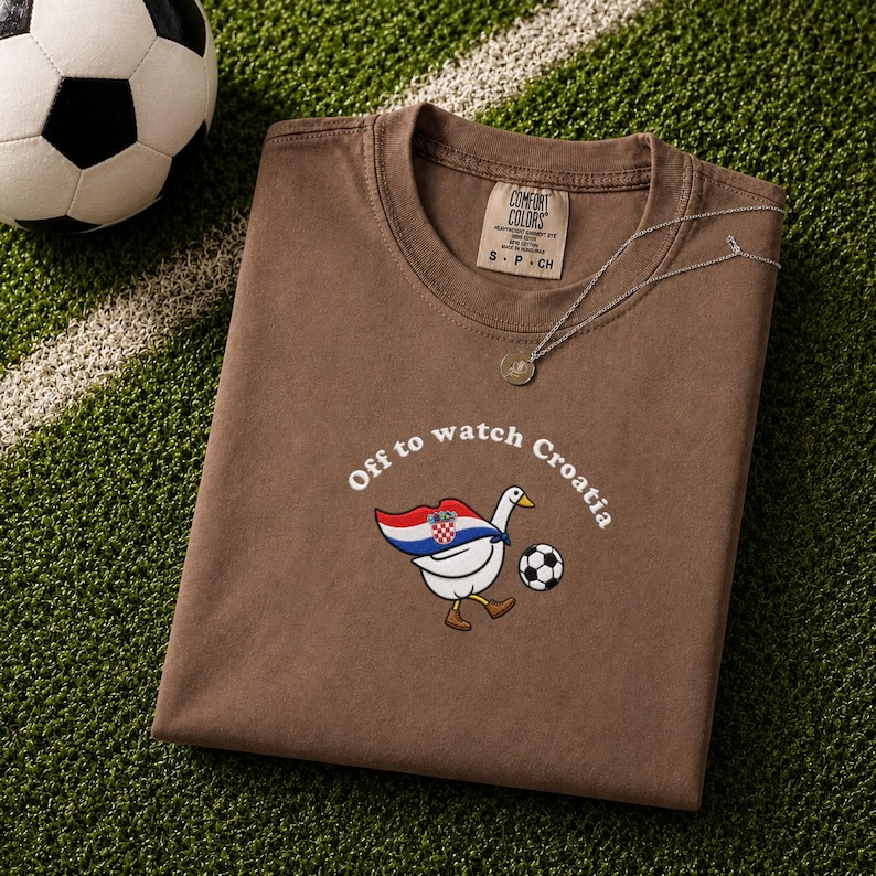 K&ouml;nnte beinhalten: Ein braunes T-Shirt mit kurzen &Auml;rmeln und einer Grafik einer wei&szlig;en Gans, die ein Cape in den Farben der kroatischen Flagge tr&auml;gt und einen Fu&szlig;ball kickt. Der Text &uuml;ber der Gans lautet "Off to watch Croatia".