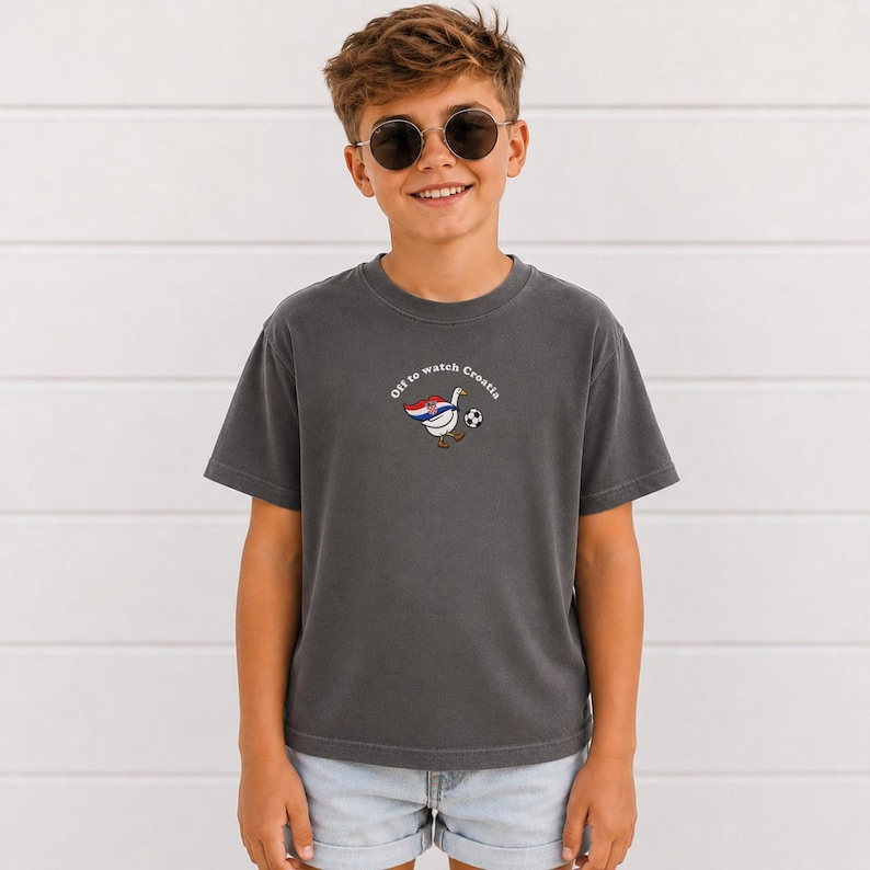 K&ouml;nnte beinhalten: Ein anthrazitfarbenes Kurzarm-T-Shirt mit einer Grafik einer wei&szlig;en Gans, die ein Cape in den Farben der kroatischen Flagge tr&auml;gt und einen Fu&szlig;ball kickt. Der Text "Off to watch Croatia" ist in einem wei&szlig;en Bogen &uuml;ber der Gans gedruckt.
