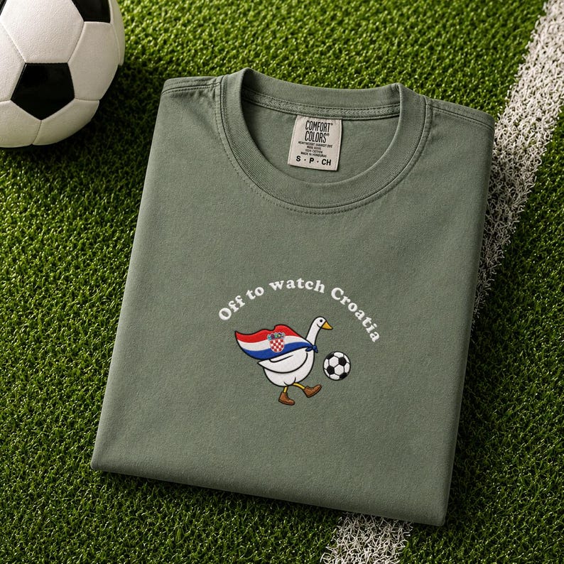 K&ouml;nnte beinhalten: Ein olivgr&uuml;nes T-Shirt mit Rundhalsausschnitt und einer Grafik einer wei&szlig;en Gans, die einen Umhang mit der kroatischen Flagge tr&auml;gt, gegen einen Fu&szlig;ball tritt und den Text "Off to watch Croatia" in wei&szlig;en, gebogenen Buchstaben zeigt.