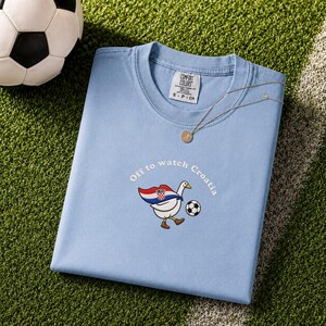 K&ouml;nnte beinhalten: Ein hellblaues T-Shirt mit Rundhalsausschnitt und einer Stickerei, die eine wei&szlig;e Gans mit einem Umhang in den Farben der kroatischen Flagge zeigt, die einen Fu&szlig;ball kickt. Der Text "Off to watch Croatia" steht dar&uuml;ber. Comfort Colors Etikett.