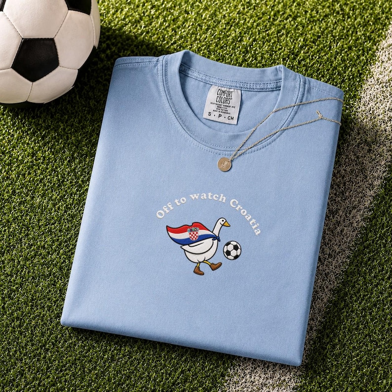 K&ouml;nnte beinhalten: Ein hellblaues T-Shirt mit Rundhalsausschnitt und einer Stickerei, die eine wei&szlig;e Gans mit einem Umhang in den Farben der kroatischen Flagge zeigt, die einen Fu&szlig;ball kickt. Der Text "Off to watch Croatia" steht dar&uuml;ber. Comfort Colors Etikett.