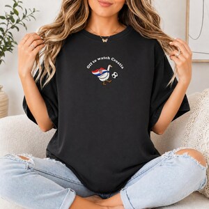 K&ouml;nnte beinhalten: Ein schwarzes Kurzarm-T-Shirt mit einer Grafik einer wei&szlig;en Gans, die ein Cape mit dem Muster der kroatischen Flagge tr&auml;gt und einen Fu&szlig;ball kickt. Der Text "Off to watch Croatia" ist in einem wei&szlig;en Bogen &uuml;ber der Gans abgebildet.