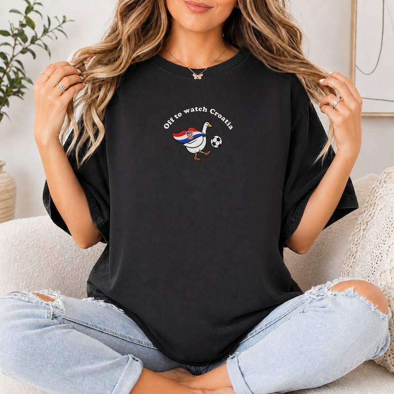 K&ouml;nnte beinhalten: Ein schwarzes Kurzarm-T-Shirt mit einer Grafik einer wei&szlig;en Gans, die ein Cape mit dem Muster der kroatischen Flagge tr&auml;gt und einen Fu&szlig;ball kickt. Der Text "Off to watch Croatia" ist in einem wei&szlig;en Bogen &uuml;ber der Gans abgebildet.
