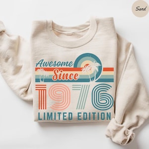 May include: A sand-colored crewneck sweatshirt featuring a graphic print with the text Awesome Since 1976 Limited Edition. The design includes retro-style horizontal stripes, palm tree silhouettes, and a large 1976 in a linear font.