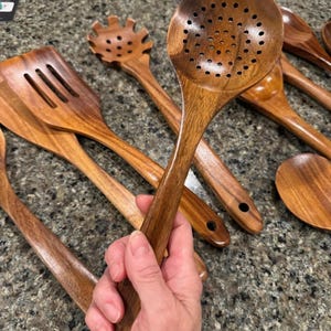 May include: A collection of wooden kitchen utensils, including a slotted spoon, spatula, and pasta server. The utensils are made of a warm-toned wood and have a smooth finish. The slotted spoon is held in a hand.