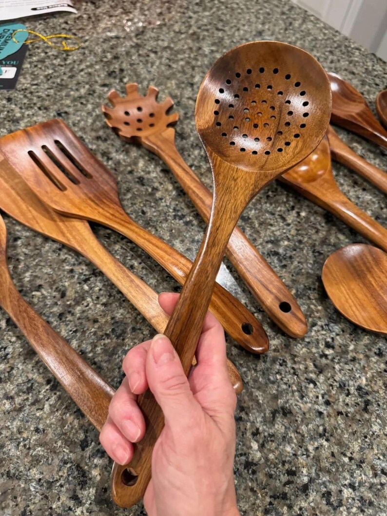 May include: A collection of wooden kitchen utensils, including a slotted spoon, spatula, and pasta server. The utensils are made of a warm-toned wood and have a smooth finish. The slotted spoon is held in a hand.