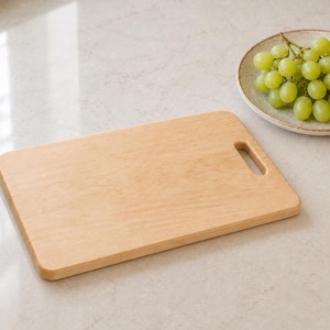 May include: A rectangular light-colored wooden cutting board with rounded corners and a single oval-shaped handle cutout on the right side. Beside the board sits a round ceramic plate filled with a cluster of fresh green grapes.