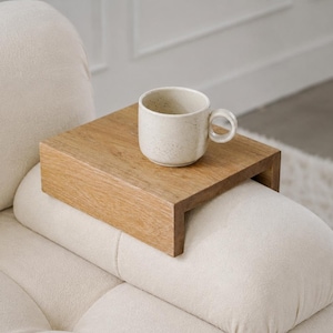 May include: A rectangular wooden armrest tray with a natural light brown wood grain finish, designed to sit securely over a sofa arm. A speckled off-white ceramic mug with a side handle is placed on the flat surface of the wooden tray.