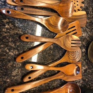 May include: A collection of wooden kitchen utensils, including spoons, spatulas, and a slotted spoon. The utensils are made of a warm-toned wood with visible grain, and each has a hole in the handle for hanging. The arrangement suggests a set for cooking.