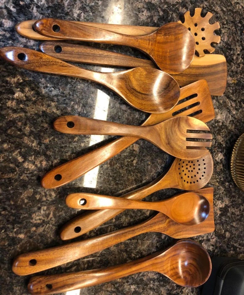May include: A collection of wooden kitchen utensils, including spoons, spatulas, and a slotted spoon. The utensils are made of a warm-toned wood with visible grain, and each has a hole in the handle for hanging. The arrangement suggests a set for cooking.