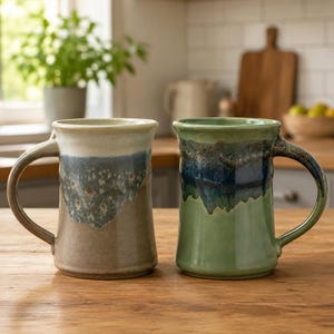 May include: Two ceramic mugs with handles, one featuring a beige base with a blue and white drip glaze at the top, and the other featuring a light green base with a dark blue and brown drip glaze at the top, both standing on a wooden surface.