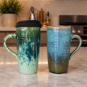 May include: Two ceramic mugs with handles. One is green and blue with a black lid, the other is blue and green. Both mugs have a textured, glazed finish. The mugs are on a marble countertop in a kitchen setting.