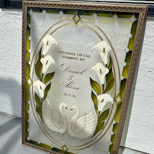 Pode incluir: Uma placa de casamento em vitral retangular numa moldura dourada com um centro oval apresentando l&iacute;rios de calla brancos e dois cisnes brancos. O texto diz: WELCOME TO THE WEDDING OF Daniel & Alexa 10.31.26. A borda tem detalhes em vidro verde.