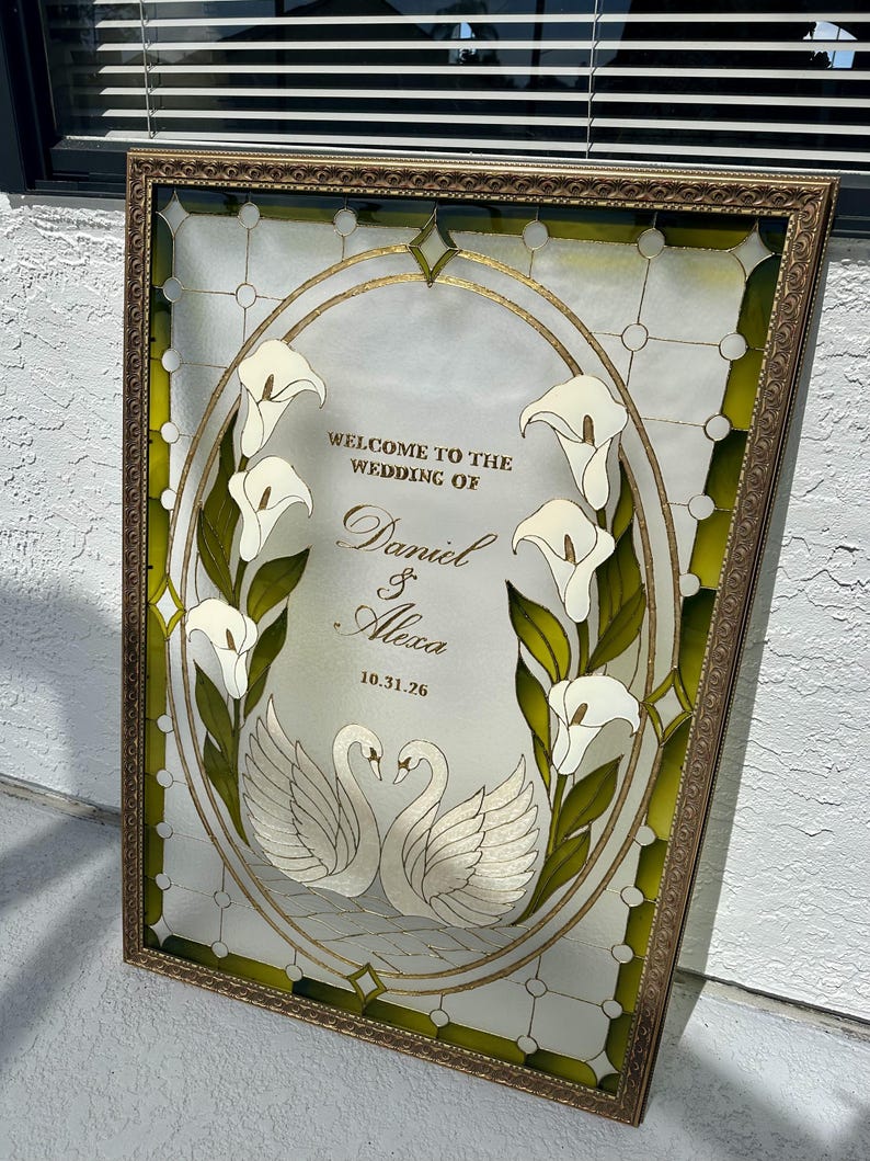Pode incluir: Uma placa de casamento em vitral retangular numa moldura dourada com um centro oval apresentando l&iacute;rios de calla brancos e dois cisnes brancos. O texto diz: WELCOME TO THE WEDDING OF Daniel & Alexa 10.31.26. A borda tem detalhes em vidro verde.