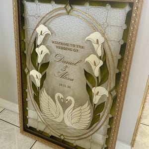 Pode incluir: Uma placa retangular emoldurada com um design em estilo vitral e um centro oval. O vidro exibe copos-de-leite brancos, dois cisnes brancos voltados um para o outro e o texto: WELCOME TO THE WEDDING OF Daniel & Alexa 10.31.26.
