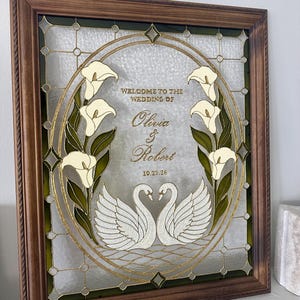 Pode incluir: Uma moldura de madeira retangular contendo um painel de vidro decorativo com um design oval central. O vidro apresenta dois cisnes brancos, copos-de-leite e o texto "WELCOME TO THE WEDDING OF Olivia & Robert 10.23.26" em letras douradas.