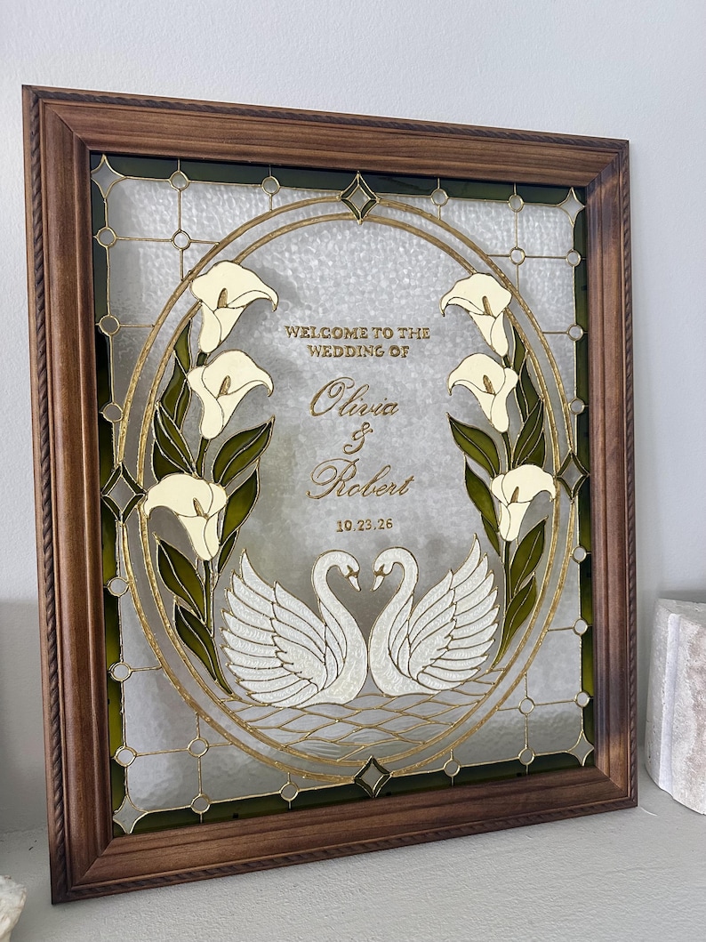 Pode incluir: Uma moldura de madeira retangular contendo um painel de vidro decorativo com um design oval central. O vidro apresenta dois cisnes brancos, copos-de-leite e o texto "WELCOME TO THE WEDDING OF Olivia & Robert 10.23.26" em letras douradas.