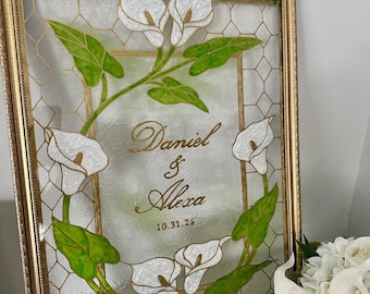 Pode incluir: Uma moldura retangular dourada contendo um painel de vidro com um padrão de favo de mel. O centro apresenta o texto dourado Daniel & Alexa e a data 10.31.26, rodeados por copos-de-leite brancos e folhas verdes pintados à mão.