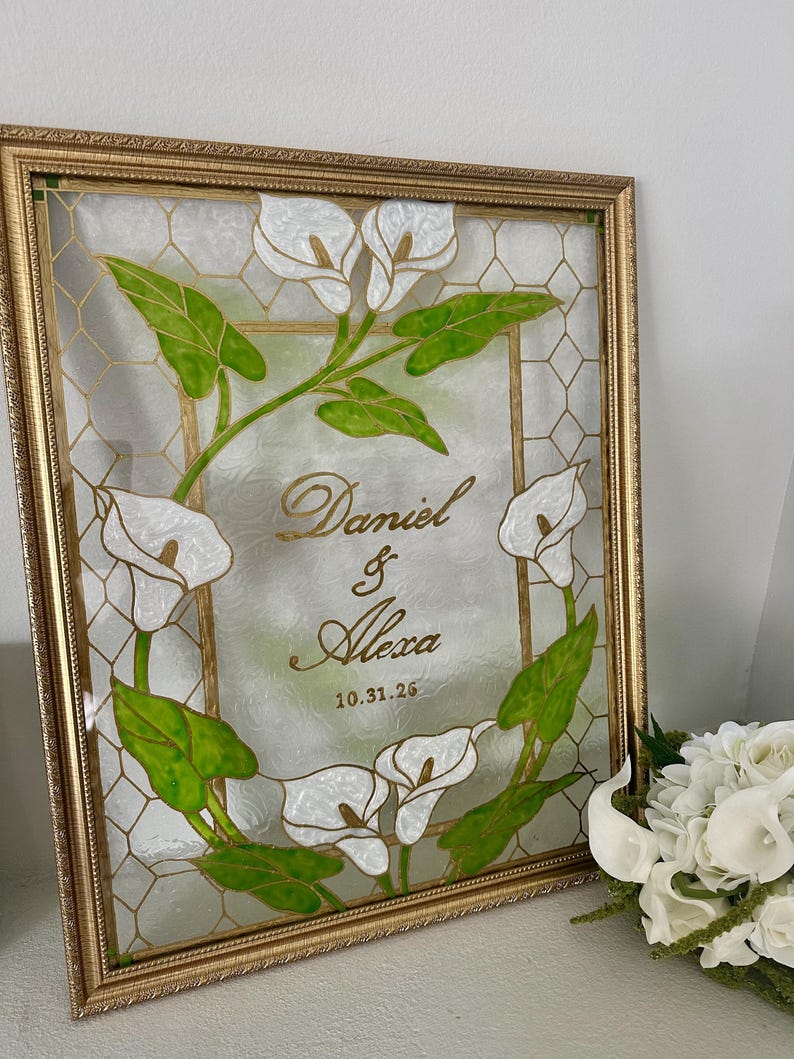 Puede incluir: Un marco rectangular dorado que contiene un panel de vidrio con un patr&oacute;n de panal. El centro presenta el texto dorado Daniel & Alexa y la fecha 10.31.26, rodeados de calas blancas y hojas verdes pintadas a mano.
