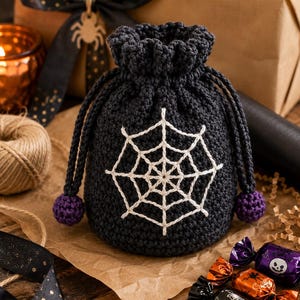 May include: A dark grey crochet drawstring pouch featuring a white embroidered spiderweb design on the front. The bag has a ruffled top edge and two long drawstrings finished with small, round, purple crocheted pom-poms at the ends.