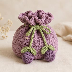 May include: A lavender crochet drawstring pouch featuring a ruffled top edge and a light green cord tie. The pouch is decorated with two small, dark purple crocheted berry clusters, each attached to the drawstring with light green leaves.
