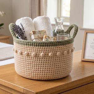 May include: A round, beige crocheted basket with a dark green rim and a row of small bobble accents. The basket contains two rolled white towels, a glass carafe, a clear glass, two wrapped soap bars, and a sprig of dried lavender.