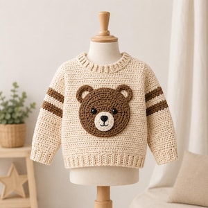 Könnte beinhalten: Ein cremefarbener, langärmeliger Häkelpullover mit einer großen, braunen Bärengesicht-Applikation auf der Brustmitte. Der Pullover hat einen gerippten Rundhalsausschnitt, Bündchen und Saum sowie zwei horizontale braune Streifen an jedem Ärmel und schwarze Knopfaugen.