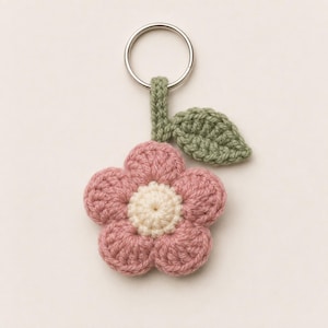 May include: A crochet flower keychain featuring five dusty pink petals surrounding a cream-colored center. A small green crocheted leaf is attached to the stem, which connects to a circular silver metal split ring for holding keys securely.