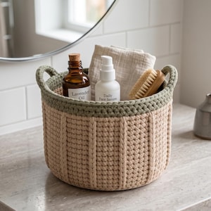 May include: A round, beige crocheted basket with a sage green rim and two integrated handles. The basket contains a brown glass bottle labeled Lavender Oil, a white pump bottle labeled Daily Face Serum, a folded beige cloth, and a wooden brush.