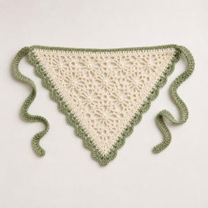 Peut inclure: Un bandana triangulaire en crochet présentant un motif de dentelle florale complexe de couleur crème. Les bords sont finis avec une bordure festonnée en fil vert sauge, et deux longs liens en crochet assortis partent des coins supérieurs.