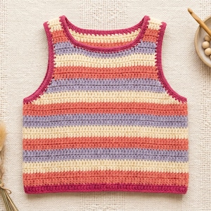 May include: A handmade crochet top in a striped pattern of coral, lavender, and cream. The top has a magenta trim and is displayed on a textured, off-white surface. A small bouquet of dried flowers and a bowl with crochet supplies are also present.