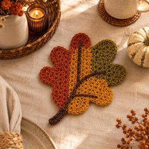 May include: A crochet leaf coaster featuring a color-blocked design in shades of burnt orange, golden yellow, and olive green. The leaf shape is defined by a dark brown crocheted stem and central vein detail, crafted with a textured yarn stitch pattern.