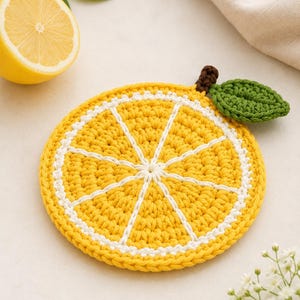Può includere: Un sottobicchiere rotondo all'uncinetto che riproduce una fetta di limone. L'oggetto presenta un centro giallo brillante con linee radiali bianche, un bordo bianco e un piccolo stelo marrone attaccato a una foglia verde all'uncinetto sul bordo.