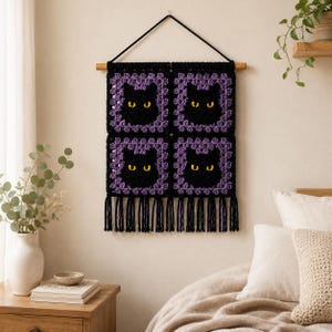 May include: A square crochet wall hanging featuring four black cat faces with yellow eyes, framed by purple borders. The piece is suspended from a wooden dowel with a black cord and finished with long black and purple fringe at the bottom edge.