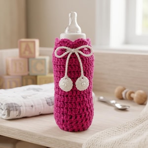 May include: A hot pink crocheted baby bottle cover with a white bow and two white pom-poms. The bottle cover is designed to fit a standard baby bottle. The image also includes a baby bottle, a knitted bunny, and baby blocks.