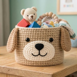 May include: A tan crocheted storage basket shaped like a dog with floppy ears, a cream-colored muzzle, and black embroidered features. The basket is filled with various small toys, including a crocheted teddy bear and wooden blocks.
