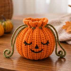 May include: A round, orange crochet pumpkin pouch with a drawstring closure. The pumpkin features a stitched black face with closed eyes and a small triangular nose. Two green crocheted cords hang from the top, ending in curled ends.