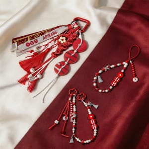 Op de afbeelding: Een verzameling rode en witte sleutelhangers en accessoires. Eén sleutelhanger heeft het woord "ARIRANG" op een lint. Andere items zijn kralenarmbanden en decoratieve elementen met kwastjes en bloemaccenten.