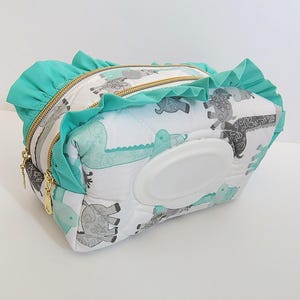 May include: A quilted fabric pouch featuring a white background with a repeating pattern of gray and teal elephants. The pouch has a teal ruffled trim, gold-toned metal zippers, and a white plastic flip-top opening centered on the front panel.