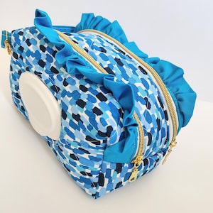 May include: A blue and white patterned baby wipe dispenser with a white lid. The bag has a quilted design with blue ruffles and gold zippers. The bag is designed to hold baby wipes.