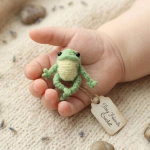 May include: A small, crocheted frog in shades of green and cream, held in a hand. The frog has black eyes and a tag that reads "Tiny Friends Crochet."